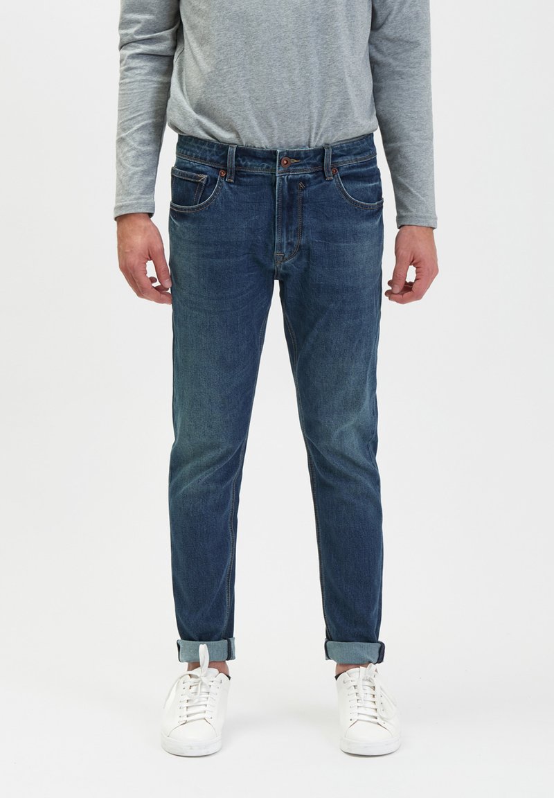 Niebieskie dżinsy w slim fit, z pięcioma kieszeniami, z lekkim wyblaknięciem i podwiniętymi mankietami, w połączeniu z białymi sneakersami i szarym, długim rękawem.