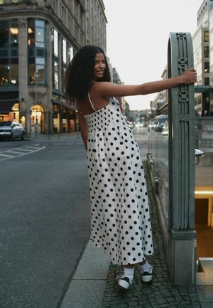 Młoda kobieta w białej sukience w grochy i trampkach opiera się o miejskie barierki o zmierzchu, uśmiechając się do kamery.