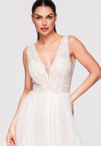 Femme aux cheveux foncés portant une robe blanche en dentelle sans manches avec un profond décolleté en V et une jupe plissée, posant avec une main sur la hanche sur un fond uni.