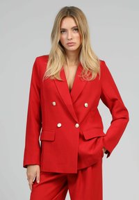 Femme blonde portant un blazer rouge à double boutonnage avec des boutons dorés et un pantalon large assorti, main dans la poche, fond neutre.