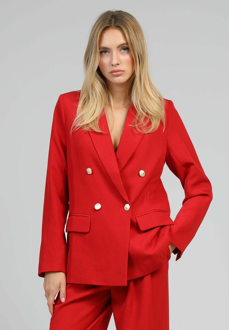 Femme blonde portant un blazer rouge à double boutonnage avec des boutons dorés et un pantalon large assorti, main dans la poche, fond neutre.