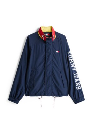 Chaqueta azul marino con un collar rojo y amarillo, puños elásticos, dos bolsillos laterales y "TOMMY JEANS" en blanco en la manga. Tejido ligero.