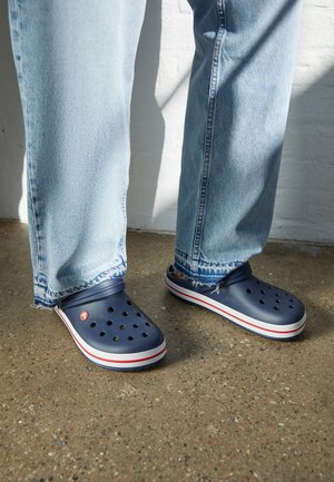 Marineblaue Clogs mit Belüftungslöchern, weißen und roten Akzenten an der Sohle, kombiniert mit hellblauen, kaputten Jeans, auf einer strukturierten Oberfläche.