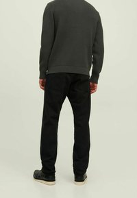 Persona in piedi con le spalle rivolte, che indossa un maglione a coste grigio scuro, pantaloni neri con tasche posteriori e scarpe nere con suole beige.