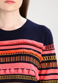 Femme portant un pull bleu marine avec des rayures multicolores horizontales, des franges et des détails en pompons sur la poitrine et les manches.