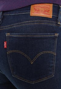 Jeans de mezclilla azul oscuro con una etiqueta de cuero que presenta gráficos en relieve. La costura amarilla resalta el diseño curvado de los bolsillos traseros.