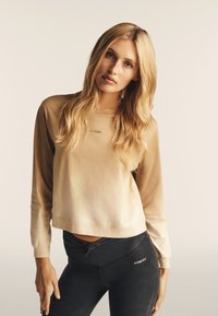 Top lungo manica color beige a taglio corto con un leggero gradiente e logo "Freddy", abbinato a leggings neri a vita alta con dettaglio nodo.