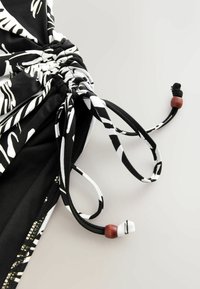 Cordon à cordonnet à motif noir et blanc avec extrémités texturées, doté d'un accent en perle de bois et d'un cordon noir. Le tissu semble lisse et léger.