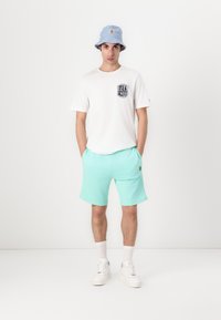 T-shirt branca com um logo no peito, calções verde menta, chapéu de balde azul claro, ténis brancos com solas texturizadas e meias até ao tornozelo.