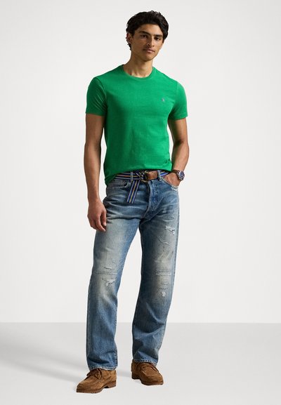 T-shirt de algodão verde, decote redondo, usado com jeans de ganga azul claro com detalhes desgastados, sapatos de camurça castanhos e um cinto multicolorido.