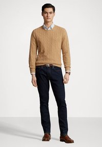 Pulover tricotat cu model de cablu de culoare bej, purtat peste o cămașă cu guler de culoare albastru deschis, asortat cu blugi de culoare albastru închis și pantofi din piele maro. Design simplu, corect ajustat.