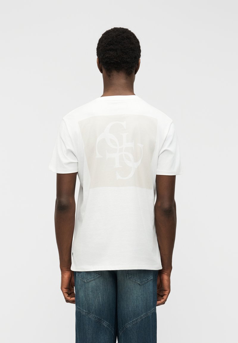 Άνδρας που φοράει λευκό κοντομάνικο μπλουζάκι με διακριτικό σχέδιο από διαπλεκόμενα γράμματα στην πλάτη, συνδυασμένο με τζιν μπλε παντελόνι, στραμμένος πλάτη.