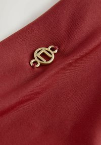 Červená tkanina s hladkou textúrou má zlatý kruhový logo akcent pripevnený marónovým povrazcom. Dizajn je minimalistický a elegantný.
