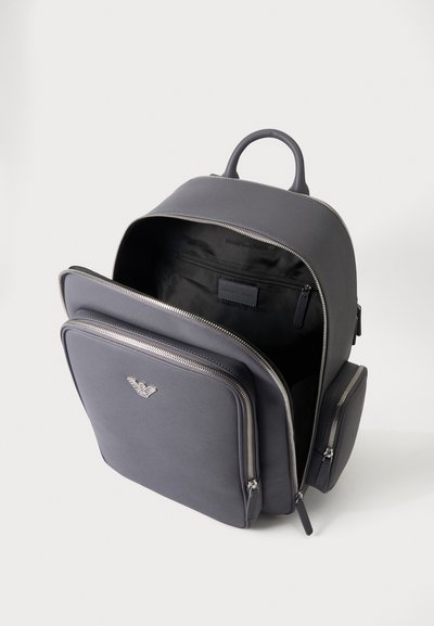 Sac à dos gris foncé avec plusieurs compartiments zippés ouverts, montrant la doublure intérieure noire et un emblème métallique frontal.