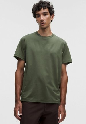 Giovane uomo con capelli ricci che indossa una maglietta verde oliva tinta unita a maniche corte e pantaloni scuri su sfondo grigio chiaro.