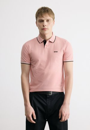 Ung mann iført en rosa polotrøye med svart kant og logo, kombinert med svarte bukser og et svart belte, stående foran en hvit bakgrunn.