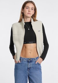Fleece-Weste in Off-White mit Reißverschluss, ausgestattet mit seitlichen Taschen und einer strukturierten Oberfläche, kombiniert mit einem schwarzen Oberteil und blauen Jeans.
