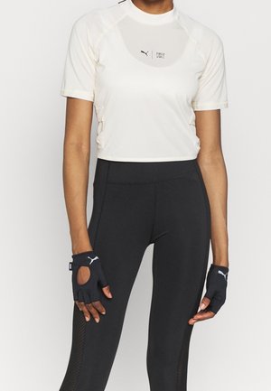 Woman wearing white Puma short-sleeve crop top, black high-waist leggings, and black fingerless Puma gloves, standing against a plain background.