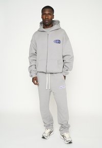 Sudadera gris con cremallera y bolsillos frontales, que presenta un logotipo azul. Combinada con pantalones de chándal a juego, ambos de suave material de forro polar, con cordón.