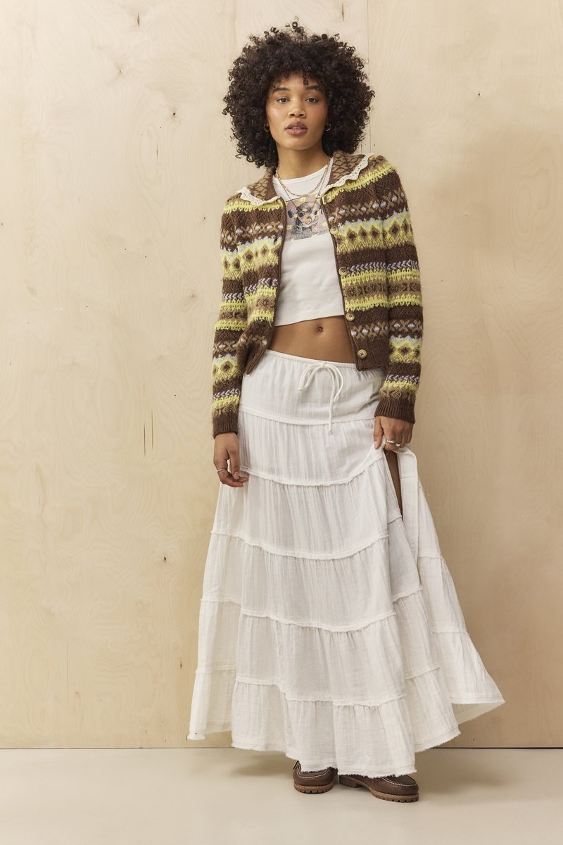 Strickjacke mit braunen, gelben und grünen Mustern, kombiniert mit einem weißen, gestuften Maxirock. Die Accessoires bestehen aus Halsketten und braunen Schuhen.