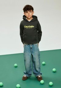 Tmavě šedá mikina s logem „Courtside“, modré volné džíny a hnědé boty. Okolo jsou zelené tenisové míčky na zeleném pozadí.