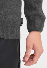 Jersey gris de punto con puños acanalados, textura de tela visible y una etiqueta de logo en el dobladillo, combinado con pantalones negros y una mano.