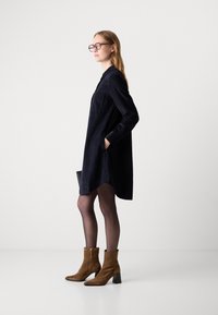 Una donna che indossa un abito camicia a maniche lunghe nero, collant neri trasparenti, stivali alla caviglia in suede marroni e occhiali, in posa di profilo su uno sfondo bianco.