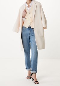 Béžový kabát přes krémovou vestu, modré džíny s ohrnutými nohavicemi a černé sandály na podpatku. Oblečení má čisté linie a kontrastní barvy.