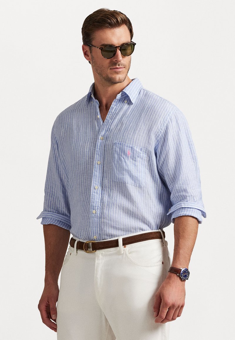 Zila un balta svītraina krekliņa ar pogām un krūšu kabatu, sarullētām piedurknēm, apvienots ar baltām biksēm un brūnu jostu. Melni saulesbrilles un pulkstenis.