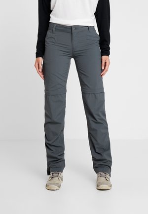 Personne portant un pantalon de randonnée convertible gris foncé à fermeture éclair, des baskets gris clair et un t-shirt à manches longues blanc et noir, debout sur un sol blanc.