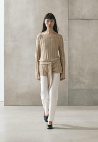 Woman walking, wearing beige ribbed sweater with waist tie, white pants, and black ballet flats, against a plain concrete wall background.