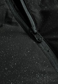 Material negru rezistent la apă, vedere de aproape cu un fermoar; suprafața prezintă picături, indicând rezistență la umezeală. Finisaj texturat, accesorii netede.