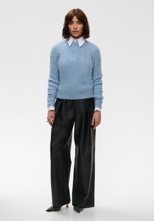 Donna con capelli castani fino alle spalle che indossa un maglione a maglia azzurro chiaro sopra una camicia bianca con colletto, pantaloni neri a gamba larga e tacchi borchiati.