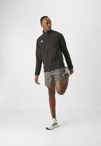 Czarna sportowa kurtka z wysokim kołnierzem, zamkiem z przodu i białym logo. Szare szorty i białe sneakersy z czarnymi akcentami. Mężczyzna rozciąga nogę.