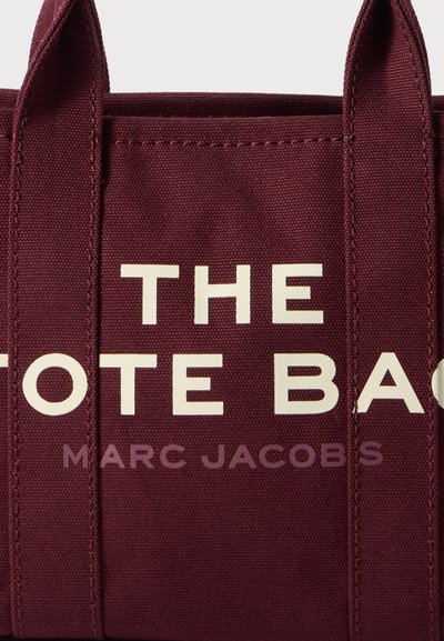 Sac fourre-tout bordeaux en matériau durable, avec de grandes lettres blanches : "LE SAC FOURRE-TOUT" et "MARC JACOBS" à l'avant.