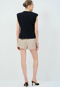 Top nero senza maniche con design strutturato sulle spalle, abbinato a pantaloni corti beige chiaro su misura e mule nere con tacco alto. Tessuto dalla texture liscia.