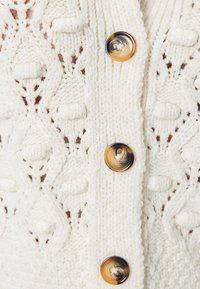 Weißer, gestrickter Cardigan mit strukturierten Mustern und großen, gewellten braunen Knöpfen. Die Nahaufnahme zeigt die Details und die Verarbeitung des Stoffes.