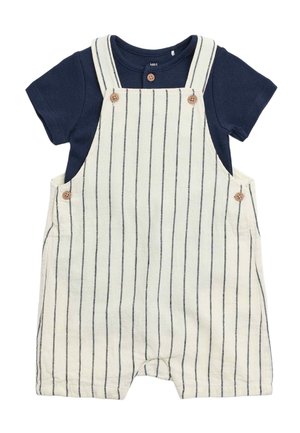 Camicia blu navy a maniche corte sotto pantaloncini in lino bianco con righe verticali blu navy e bottoni marroni sulle bretelle e sui lati.