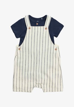 Camicia blu navy a maniche corte sotto pantaloncini in lino bianco con righe verticali blu navy e bottoni marroni sulle bretelle e sui lati.