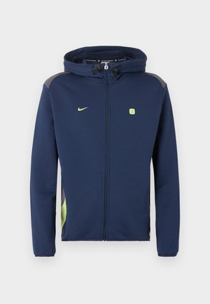 Μπλε ναυτικό μπουφάν Nike με φερμουάρ ολόκληρο, κουκούλα, ανοιχτοπράσινο λογότυπο και λεπτομέρειες, και κορδόνια στην κουκούλα πάνω σε μονόχρωμο φόντο.