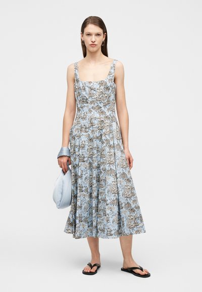 Μια γυναίκα στέκεται φορώντας ένα χωρίς μανίκια, μπλε φόρεμα με φλοράλ σχέδιο μέχρι το μέσο της γάμπας, κρατώντας μια ανοιχτό μπλε τσάντα και φοράει μαύρες σαγιονάρες με κρίκους στα σκουλαρίκια.