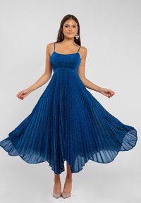 Nainen wear sleeveless, pleated, sparkly blue midi dress, joka pitää sivuja ulkona, yhdistettynä roikkuviin korvakoruihin ja kimalteleviin beige korkokenkiin.