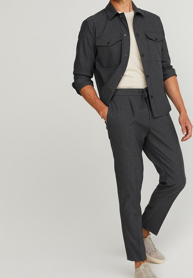 Grijze gestreepte blazer met twee borstzakken, gecombineerd met bijpassende broek. Gelaagde stof, slim fit en beige sneakers complementeren de outfit.