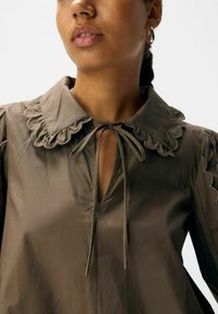 Braune Bluse mit einem wellenförmigen Kragen und Bindedetails, mit puffartigen Ärmeln und V-Ausschnitt. Glatte Textur mit subtiler Metallic-Optik.