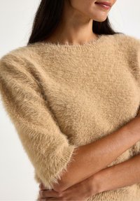Beige fluffig tröja med korta ärmar, med en mjuk textur och en vid halsringning. Armarna är korsade med slät hud synlig.