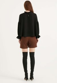 Top nero a maniche lunghe con scollo a volant, abbinato a pantaloni corti marroni e stivali neri al ginocchio, caratterizzato da una silhouette aderente e una texture liscia.