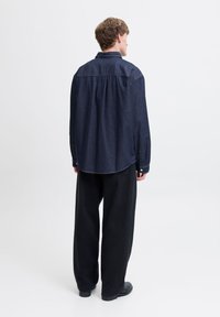 Mørk denimskjorte med avslappet passform, knappe mansjetter og buet kant, kombinert med svarte vide bukser og mørkt fottøy.