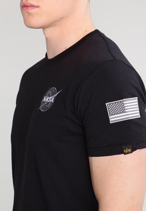 Mann trägt ein schwarzes NASA-T-Shirt mit weißem NASA-Logo auf der Brust und einem einfarbigen amerikanischen Flaggen-Patch am Ärmel vor einfachem Hintergrund.
