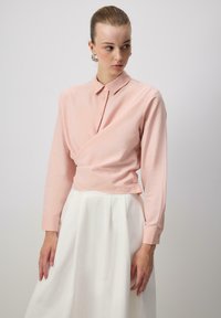 Light pink wrap shirt with a collar and long sleeves, made from a smooth fabric, paired with a flowing white skirt.