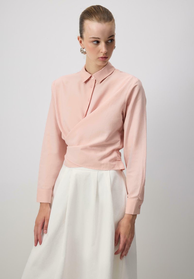 Light pink wrap shirt with a collar and long sleeves, made from a smooth fabric, paired with a flowing white skirt.